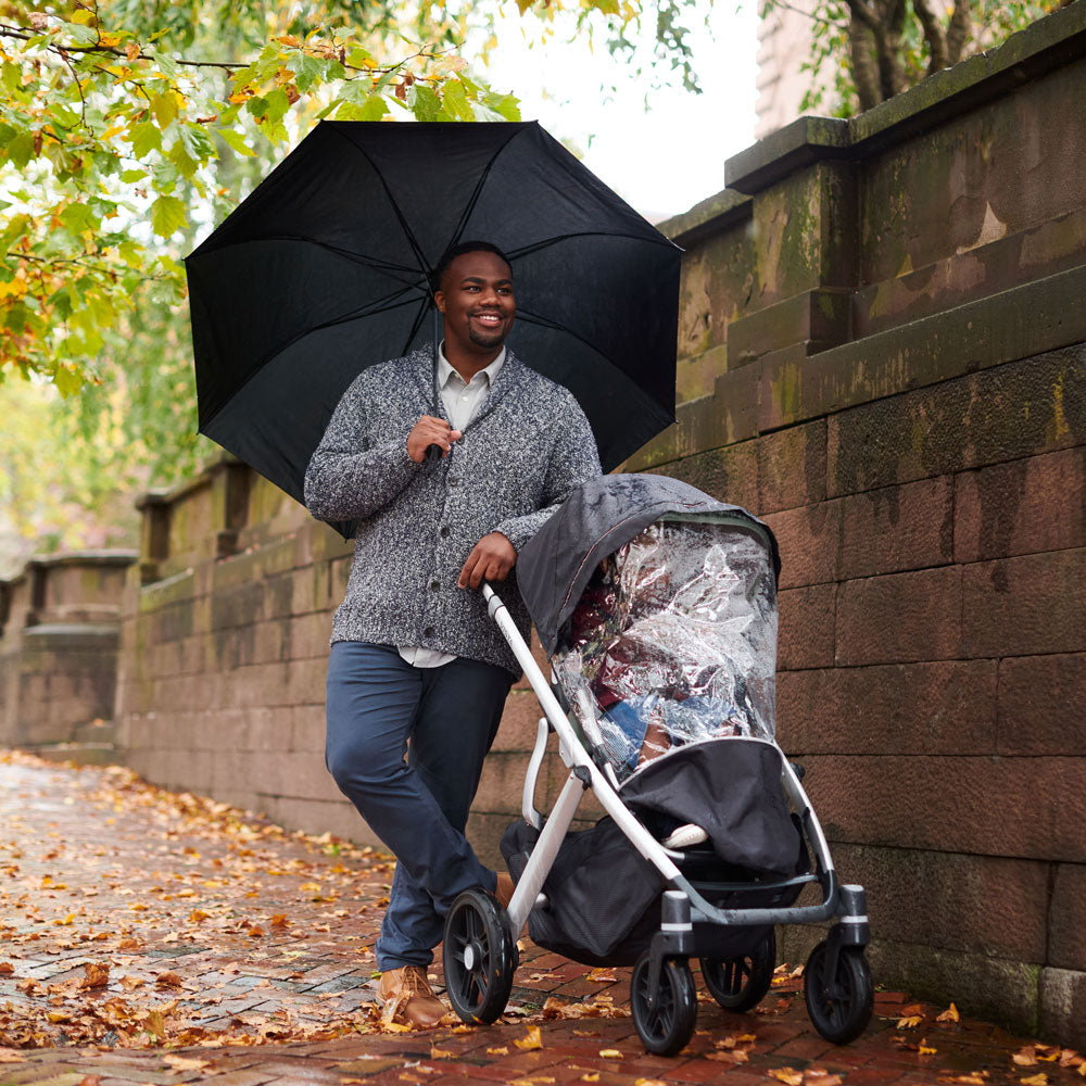 Dad stands next to child in VISTA V2 stroller with UPPAbaby VISTA / VISTA V2 / CRUZ / CRUZ V2 Performance Rain Shield attached as it rains