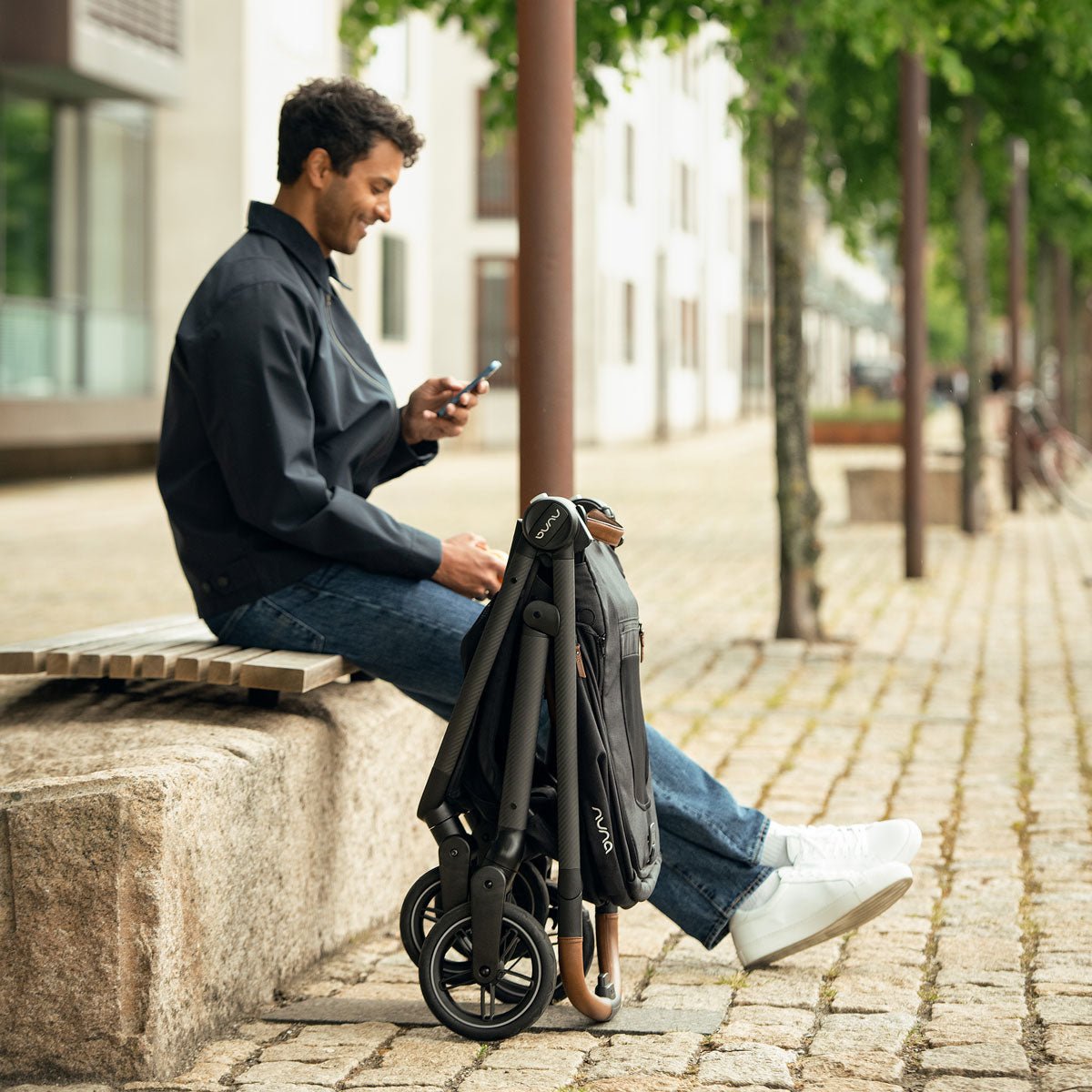 Man looks at cellphone while sitting next to folded Nuna SWIV Stroller - Caviar